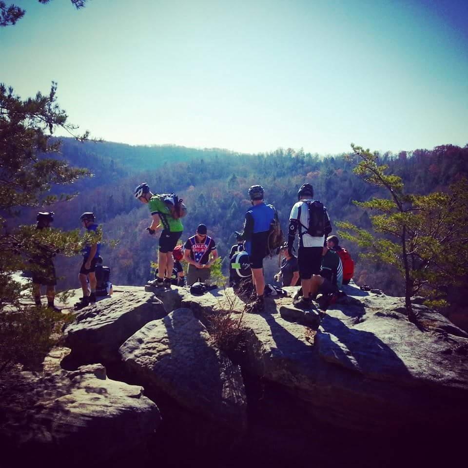A group of mountain bikers gathered on a rocky overlook, enjoying a scenic view of a forested valley beneath a clear blue sky. Some are adjusting their gear while others engage with each other, surrounded by nature. Big South Fork mountain bike trail.