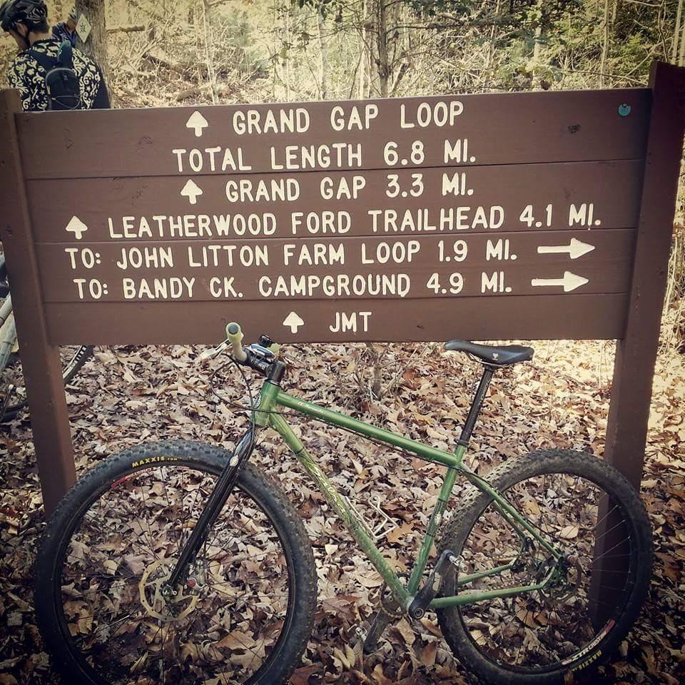 A mountain bike leaning against a trail sign in a forested area, displaying directional arrows and distances to various trails, including Grand Gap Loop, Leatherwood Ford Trailhead, John Litton Farm Loop, and Bandy Creek Campground. Leafy ground cover is visible in the surroundings. Big South Fork mountain bike trail.