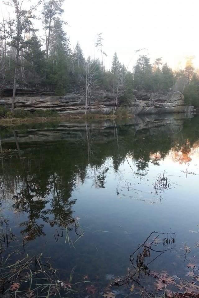 A serene landscape featuring a calm body of water reflecting tall trees and rocky cliffs. The scene captures the tranquility of nature, with a mix of greenery and autumn leaves visible near the water's edge. Big South Fork mountain bike trail.