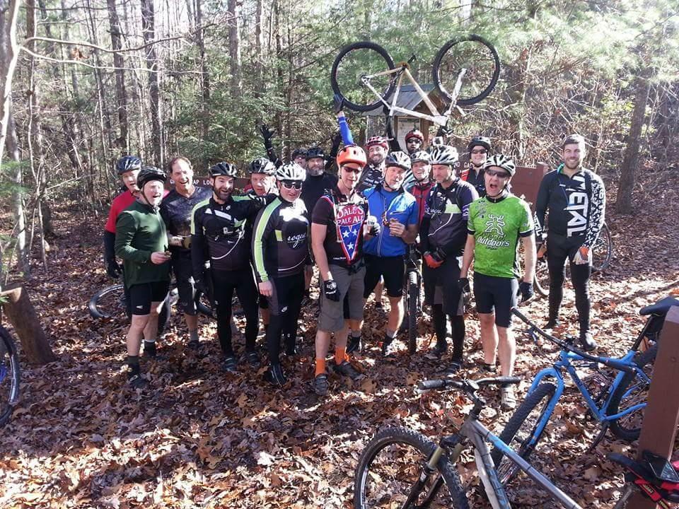 A group of mountain bikers posing together in a forested area, surrounded by fallen leaves. They are wearing various cycling attire and helmets, with some holding bikes. A few members are playfully lifting a bike overhead. In the background, there is a wooden sign partially obscured by trees. Big South Fork mountain bike trail.