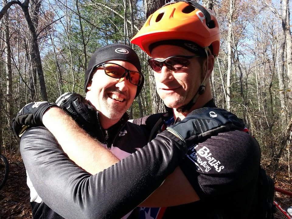 Two male mountain bikers embrace in a forest setting, both wearing sunglasses and biking gear. One biker wears an orange helmet and sports gloves, while the other has a black hat and gloves. Sunlight filters through the trees, highlighting the natural environment around them. Big South Fork mountain bike trail.