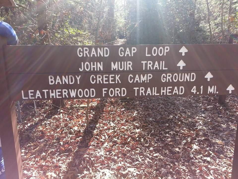 Signpost indicating trail directions in a forest, with routes for Grand Gap Loop, John Muir Trail, Bandy Creek Camp Ground, and Leatherwood Ford Trailhead (4.1 miles). Sunlight filters through the trees, illuminating the sign and surrounding landscape covered in leaves. Big South Fork mountain bike trail.