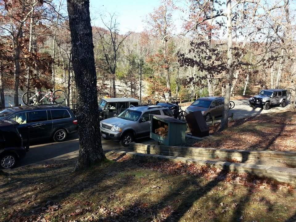 A forested area featuring several parked vehicles, including a minivan, SUVs, and a Jeep. Tall trees with autumn leaves surround the scene, capturing a peaceful outdoor atmosphere. A bicycle is visible on the roof of one vehicle, indicating potential recreational activities. A small green kiosk is partially in view, with fallen leaves scattered on the ground. Big South Fork mountain bike trail.