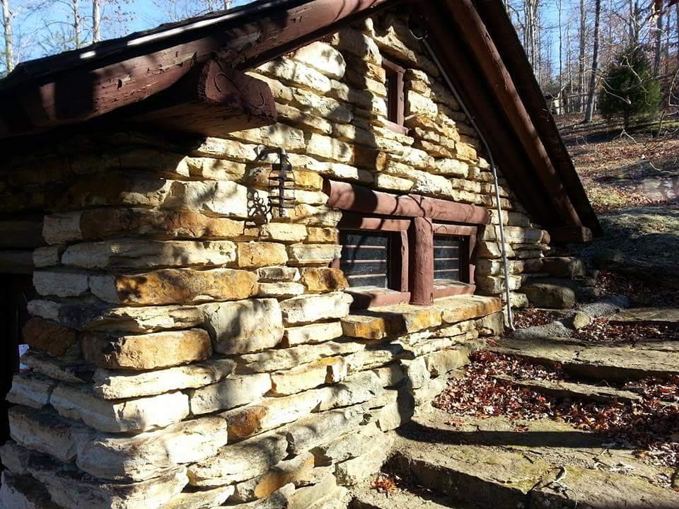 A small stone cottage with a brown wooden roof, surrounded by trees. The exterior features a rustic design with layered stone walls and wooden window frames. A set of stone steps leads up to the entrance. Sunlight casts shadows on the building, highlighting its textured surface. Big South Fork mountain bike trail.
