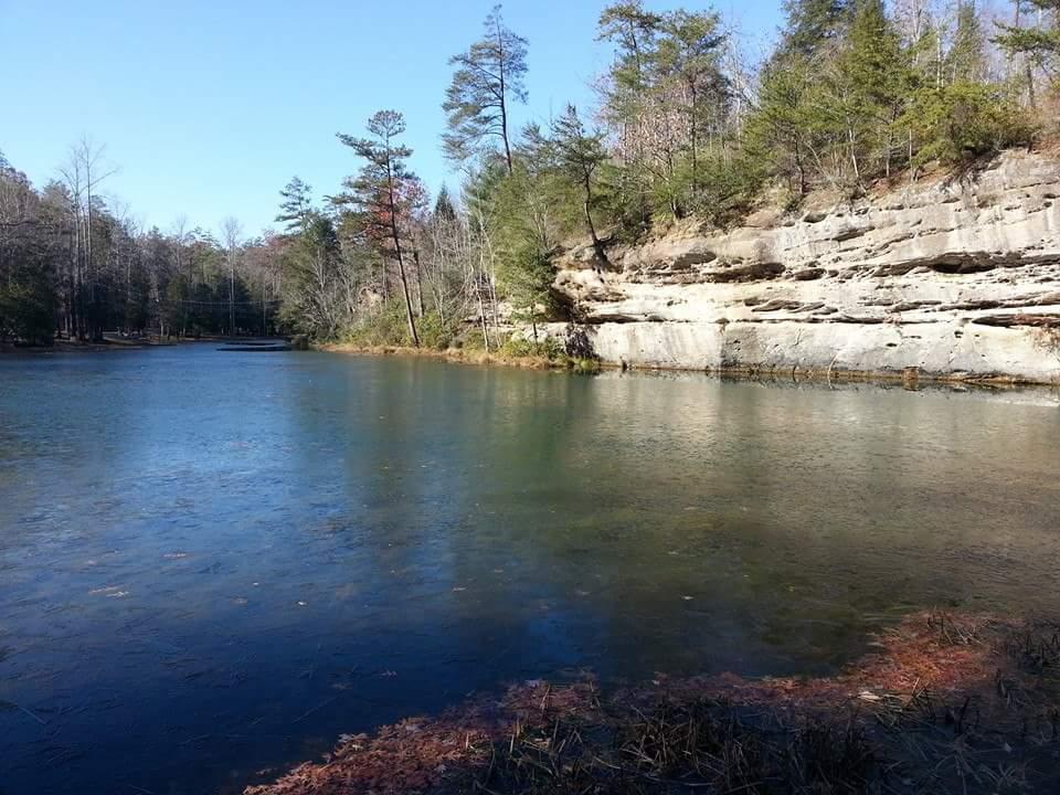 A serene river scene featuring calm water reflecting the blue sky, bordered by rocky cliffs and lush trees. The landscape showcases a mix of green pine and leafless trees, indicating a transition between seasons. Big South Fork mountain bike trail.