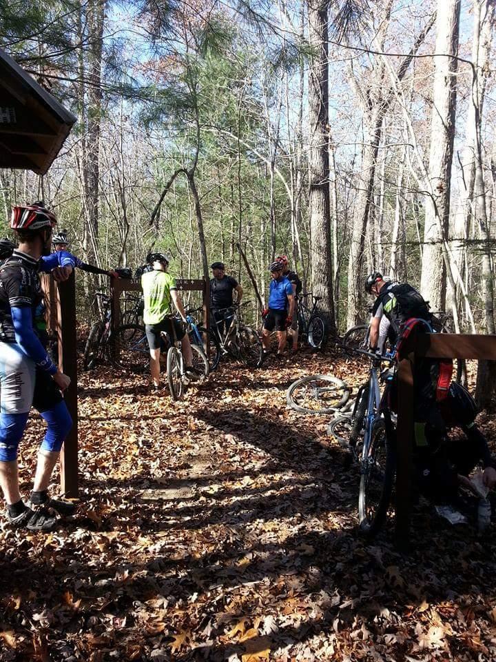 A group of mountain bikers gathered at a trailhead in a wooded area, surrounded by trees and fallen leaves. Some cyclists are adjusting their gear while others stand next to their bikes, enjoying the sunny day. The scene captures the camaraderie of outdoor biking in nature. Big South Fork mountain bike trail.
