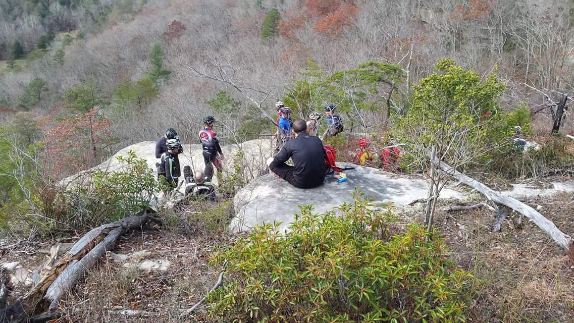 A group of mountain bikers resting on a rocky outcrop in a wooded area. The landscape features bare trees, shrubs, and distant hills. Some cyclists are sitting, while others are standing and engaging in conversation. The scene captures a moment of camaraderie and relaxation in nature. Big South Fork mountain bike trail.