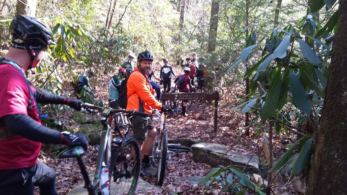 A group of mountain bikers gathered on a forest trail, with some standing by their bikes and others in the background. The setting features lush greenery, scattered leaves on the ground, and a wooden signpost visible in the foreground. The cyclists are wearing helmets and a mix of colorful riding gear. Big South Fork mountain bike trail.