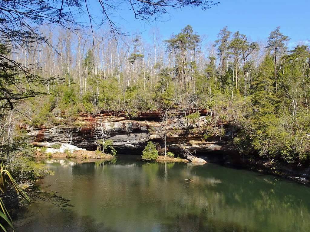 A serene landscape featuring a calm, green lake surrounded by lush trees and rocky cliffs under a clear blue sky. Big South Fork mountain bike trail.