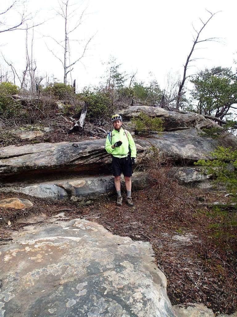A person in sports attire stands on a rocky trail surrounded by sparse vegetation and trees. The individual wears a bright yellow jacket, a helmet, and shorts, suggesting they are ready for an outdoor biking adventure. The landscape features large rock formations and a mix of greenery, indicating a natural setting. Big South Fork mountain bike trail.