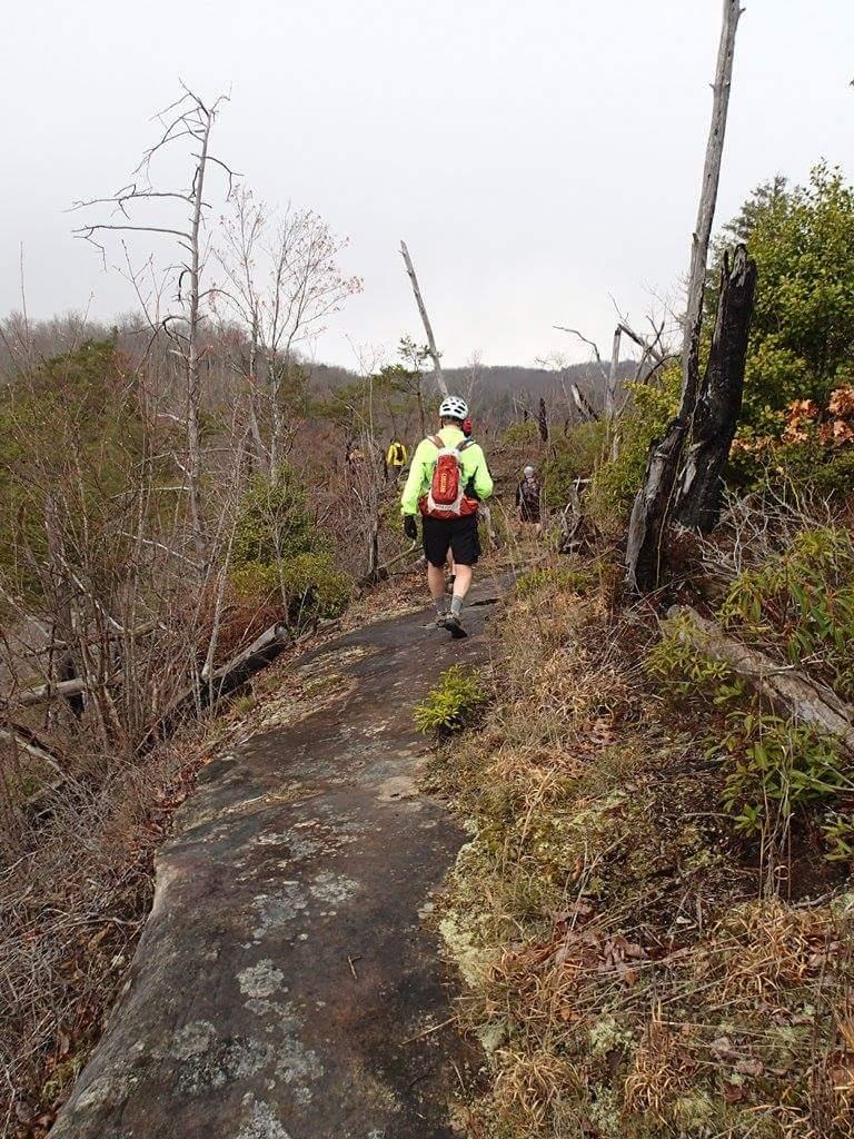 A person wearing a bright yellow jacket and a helmet is walking along a rocky trail surrounded by sparse trees and underbrush. The landscape is slightly misty, with distant hills visible in the background. Other hikers can be seen further along the path. Big South Fork mountain bike trail.