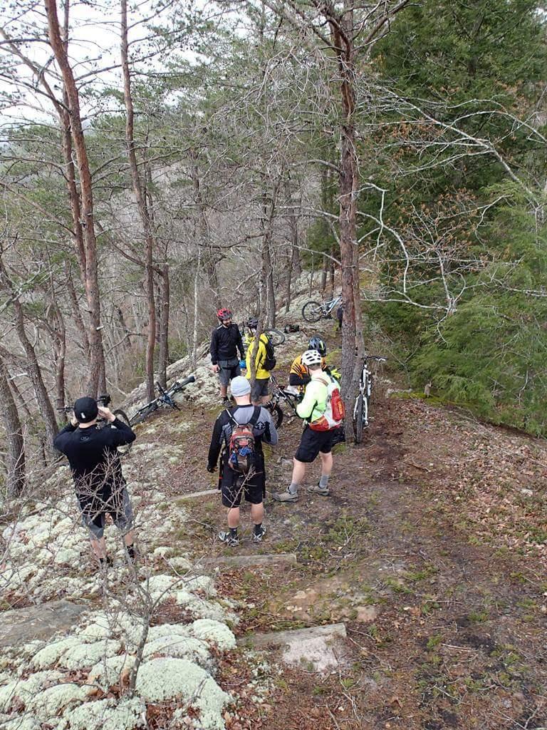 A group of mountain bikers standing on a rocky trail surrounded by trees. Some members are conversing while one person takes photos. Bicycles are parked nearby, and the scene is set in a natural, wooded area. Big South Fork mountain bike trail.
