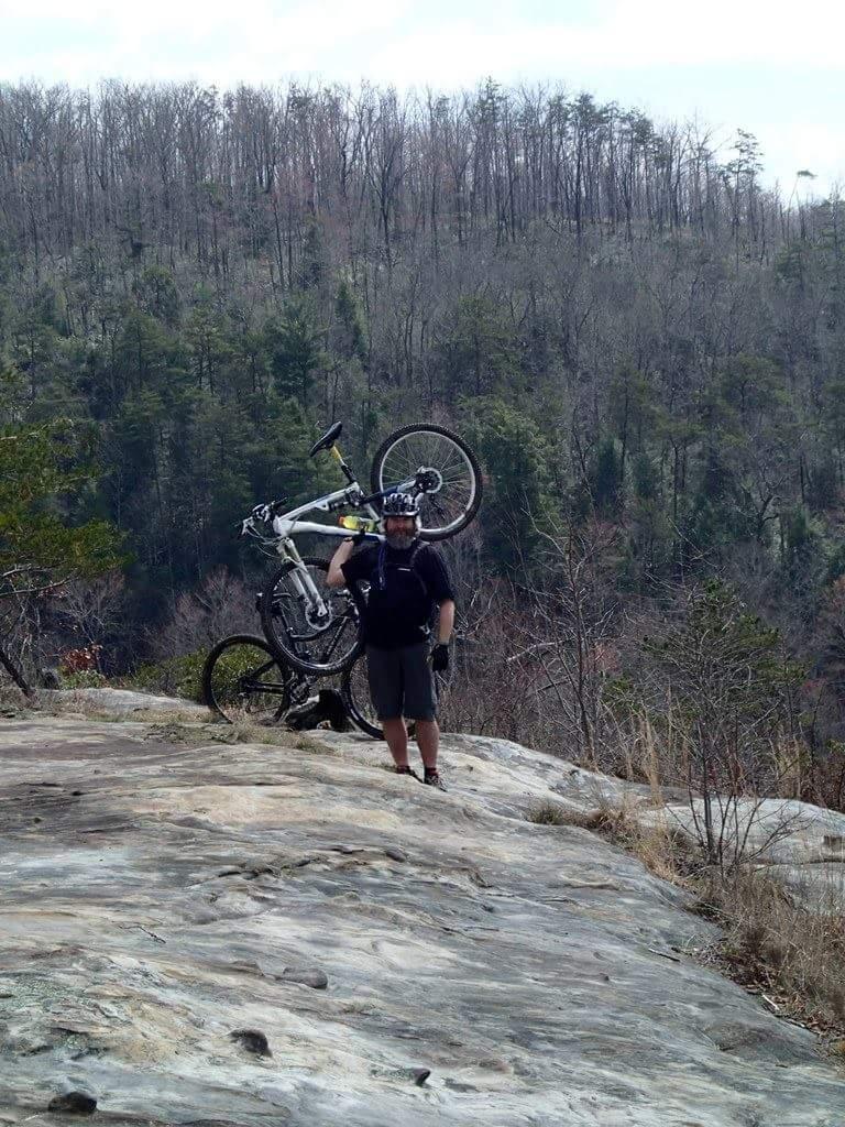 A person stands on a rocky outcrop holding a mountain bike above their head, surrounded by a forest of trees in the background. The scene captures an adventurous moment in nature, showcasing the person's enthusiasm for mountain biking. Big South Fork mountain bike trail.