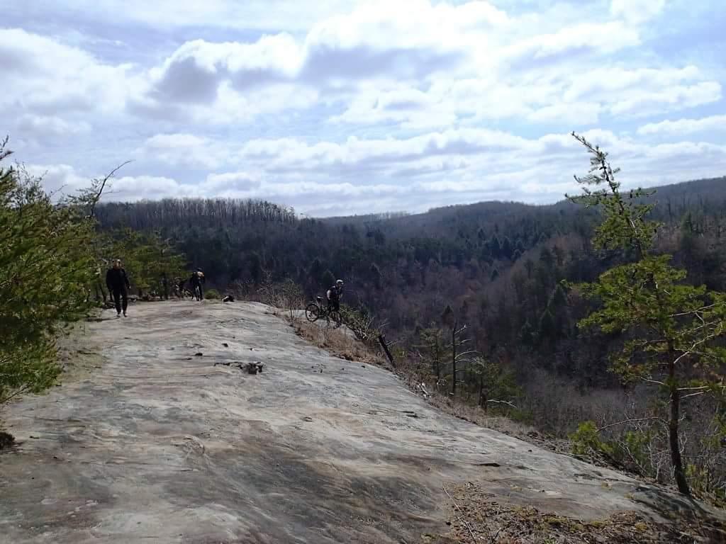 A scenic view of a rocky landscape with a few people walking and biking along the edge, surrounded by trees and rolling hills under a partly cloudy sky. Big South Fork mountain bike trail.
