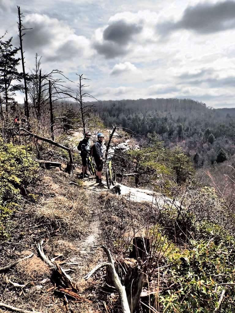 Two mountain bikers pause on a trail overlooking a lush valley, surrounded by trees and under a partly cloudy sky. The scene captures the beauty of nature and the adventure of outdoor cycling. Big South Fork mountain bike trail.