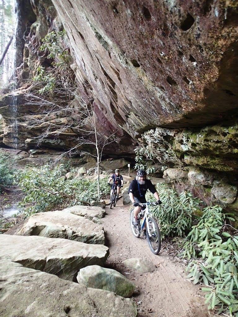 Two mountain bikers riding along a dirt trail near a rocky overhang, surrounded by greenery and boulders. The trail features a mix of dirt and stones, with a backdrop of steep rock formations and sparse trees. Big South Fork mountain bike trail.