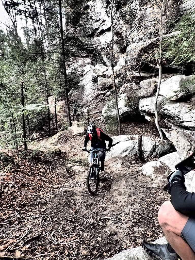 A mountain biker navigates a rocky, wooded trail surrounded by trees and large boulders, with fallen leaves covering the ground. Additional trail features can be seen in the background, creating a rugged outdoor environment. Big South Fork mountain bike trail.