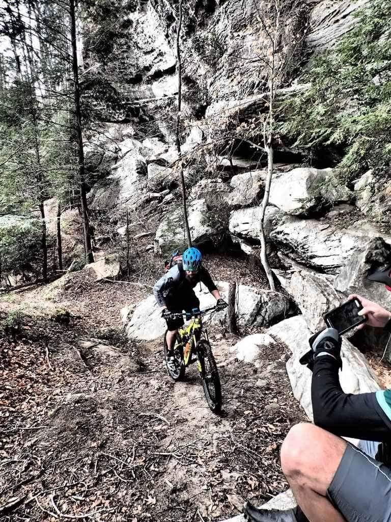A mountain biker navigates a rocky trail surrounded by trees and large boulders, while another person observes and holds a smartphone to capture the moment. Big South Fork mountain bike trail.