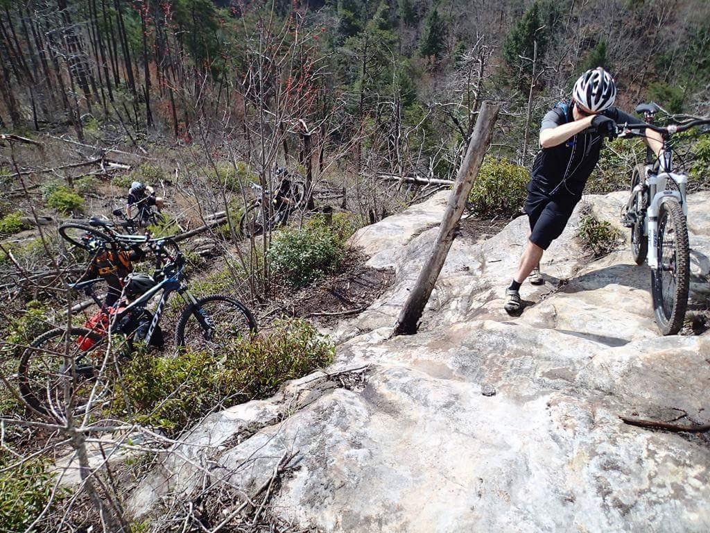 A mountain biker navigating a rocky terrain while pushing their bike uphill, with several other bikes parked nearby. The scene shows a rugged landscape with sparse vegetation, fallen trees, and a mix of greenery, indicating a remote outdoor setting. Big South Fork mountain bike trail.