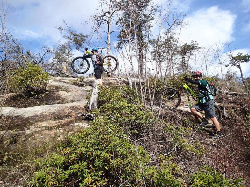 Two mountain bikers are navigating a rocky trail in a forested area. One rider is climbing uphill while carrying their bike, and the other is walking beside them, also with a bike. Surrounding them are patches of shrubs and trees under a partly cloudy sky. Big South Fork mountain bike trail.
