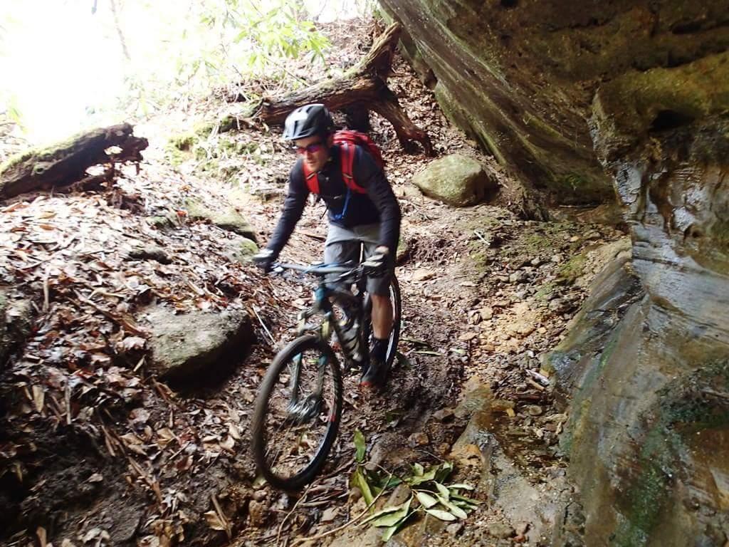 A mountain biker navigating a narrow, rocky trail surrounded by vegetation and large rock formations. The cyclist is wearing a helmet and a backpack, demonstrating an active outdoor pursuit. Big South Fork mountain bike trail.