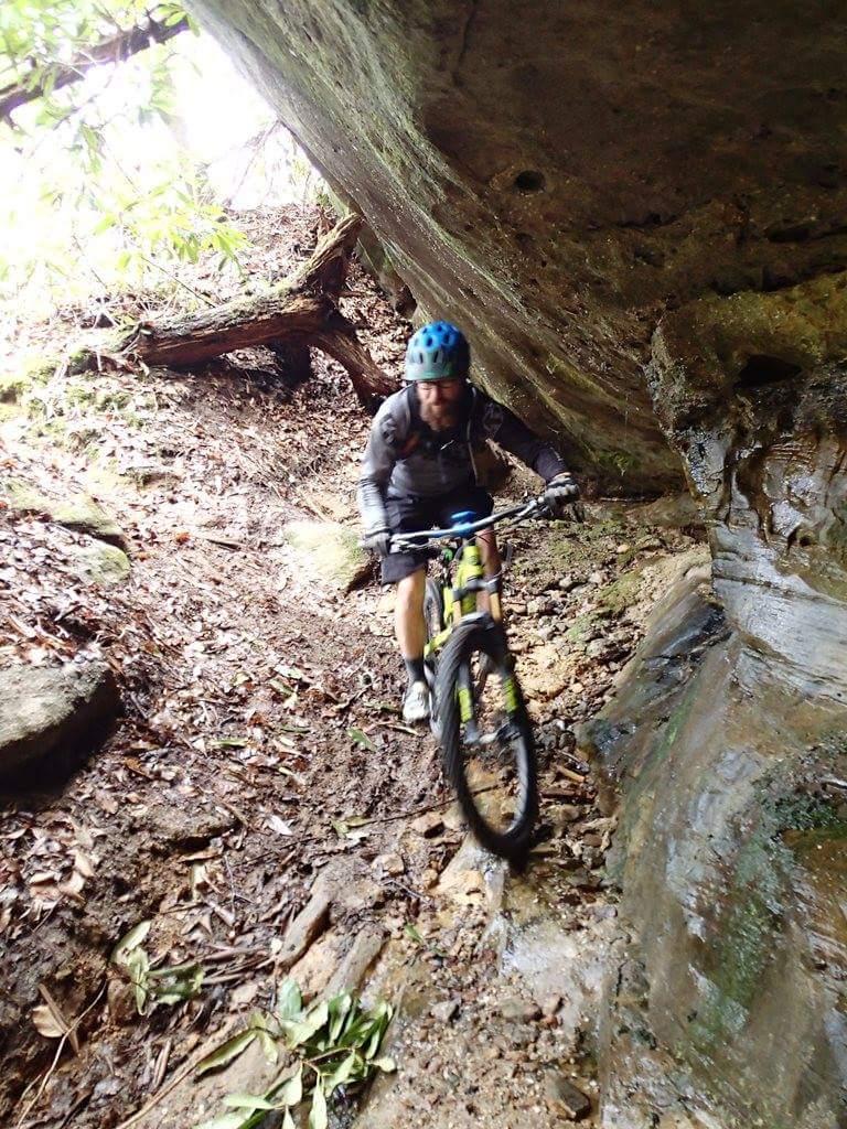 A mountain biker navigating a narrow, muddy trail under a rocky overhang surrounded by foliage. Big South Fork mountain bike trail.