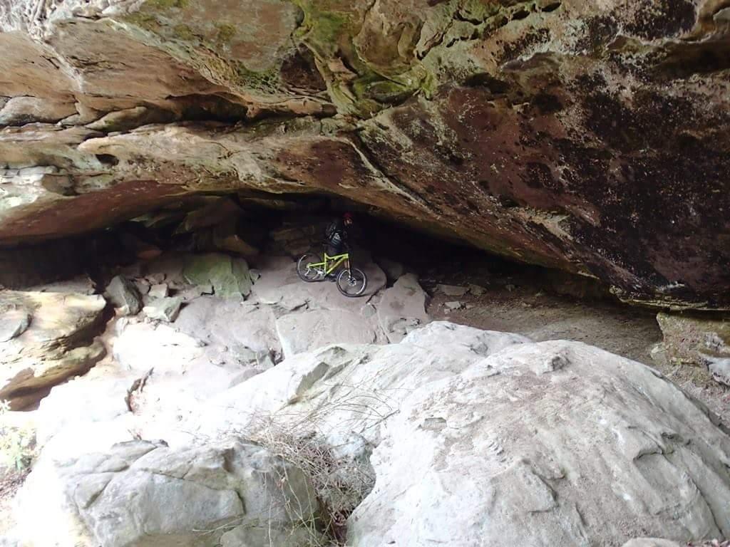 A mountain biker standing next to their bike under a large rock overhang, surrounded by rocky terrain and scattered foliage. The scene depicts a rugged natural landscape with erosion shapes visible on the rocks. Big South Fork mountain bike trail.