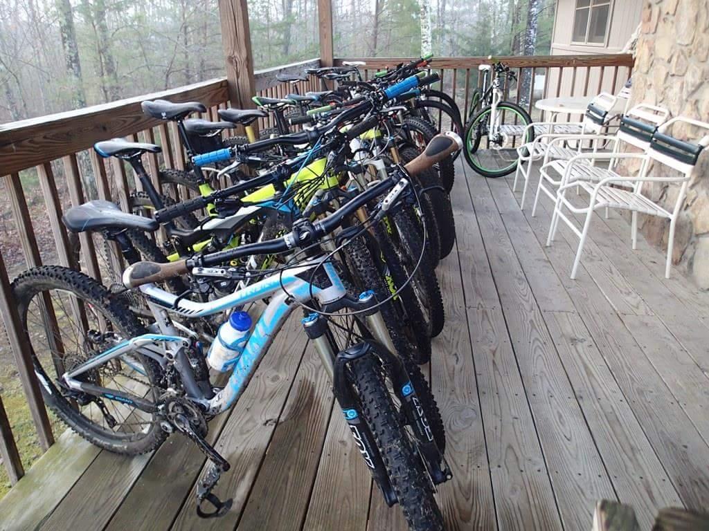 A row of mountain bikes parked on a wooden porch, with some bikes displaying colorful frames in shades of blue, green, and yellow. The background features blurred trees, indicating a natural, outdoor setting. A few plastic water bottles are visible on the bikes, and there are several white metal chairs nearby. Big South Fork mountain bike trail.