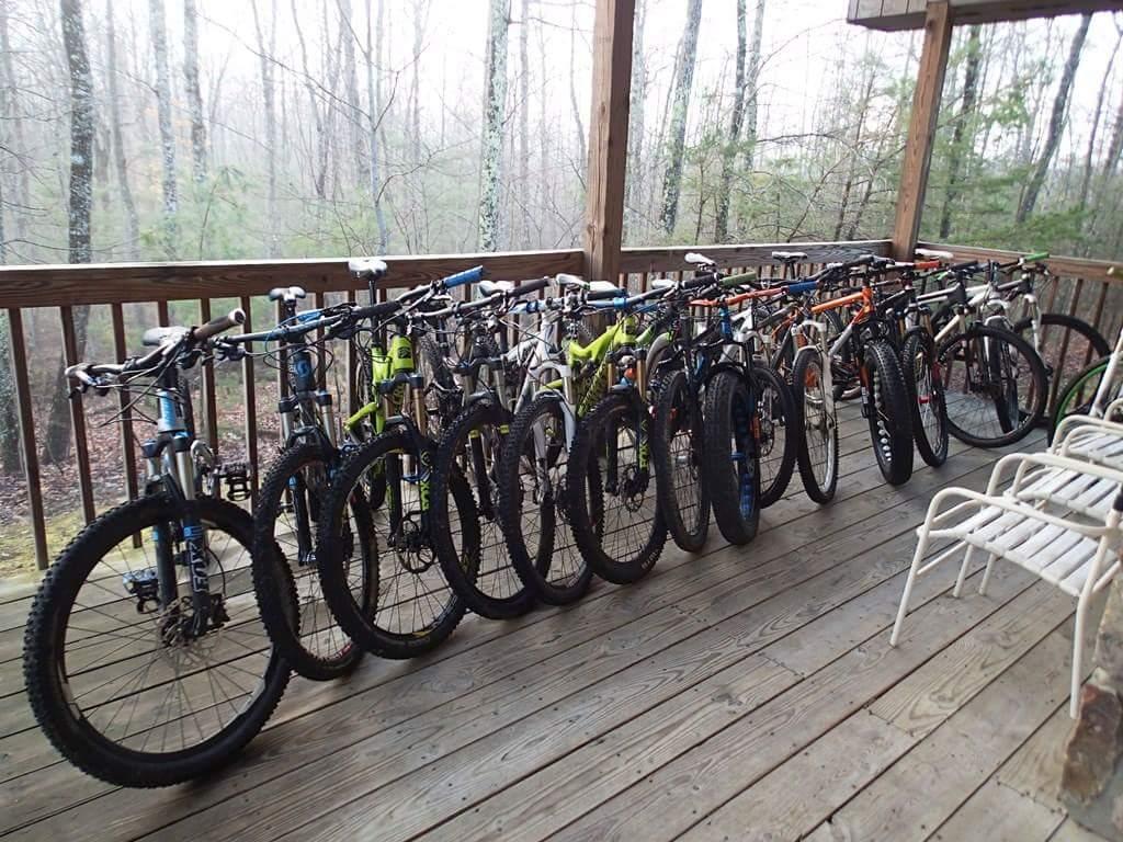 A lineup of various mountain bikes parked on a wooden deck, surrounded by a misty forest landscape in the background. Several bicycles are lined up parallel to each other, showcasing different colors and styles, while a couple of empty chairs are visible on the deck. Big South Fork mountain bike trail.
