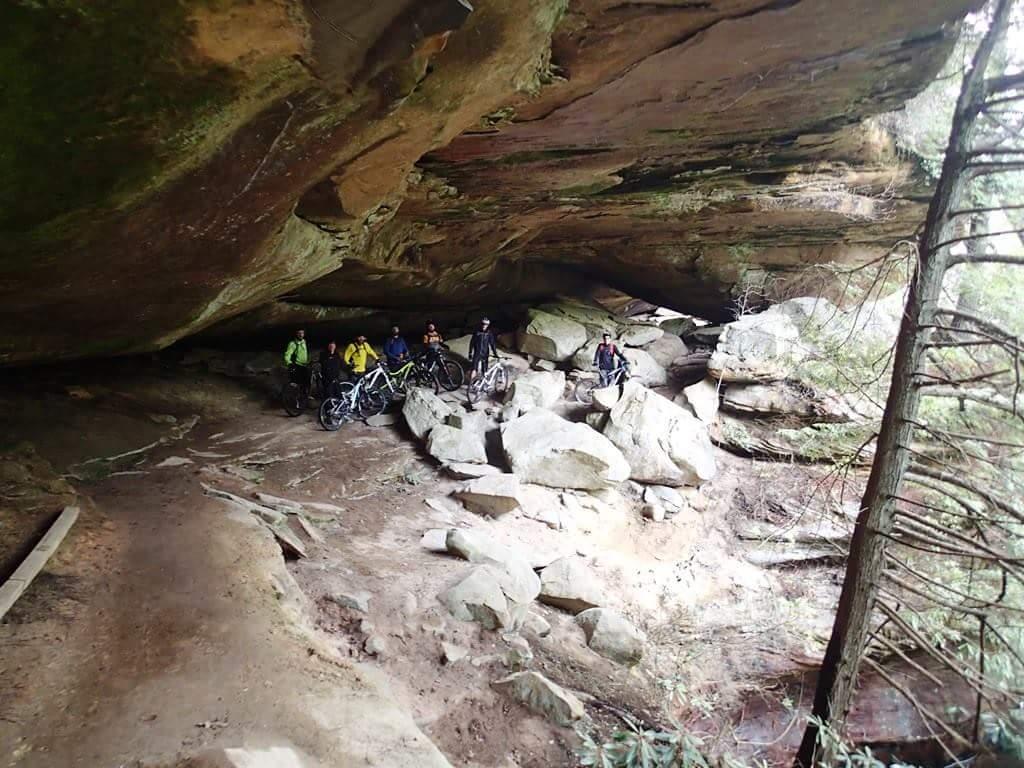 A group of mountain bikers stands under a large rock overhang, surrounded by boulders and trees in a natural setting. The trail appears rocky, indicating a rugged outdoor environment. Big South Fork mountain bike trail.