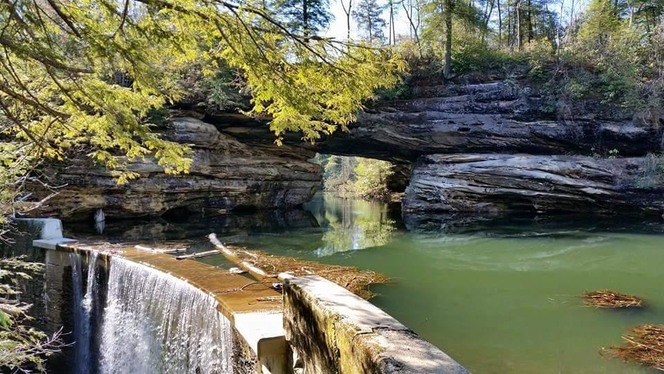 A serene landscape featuring a rocky arch over still water, with a small waterfall cascading down a concrete structure in the foreground. Lush green trees frame the scene, enhancing the natural beauty of the area. The clear water reflects the surrounding rocks and foliage, creating a tranquil atmosphere. Big South Fork mountain bike trail.