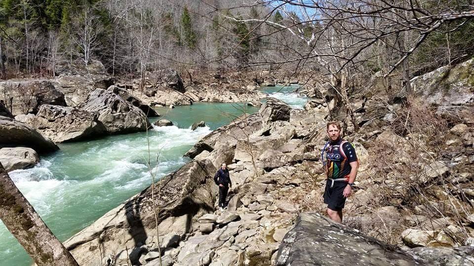 Two people hiking along a rocky riverbank with a flowing green river in the background. The scene features a mix of bare trees and evergreen vegetation on the surrounding hills, showcasing a natural landscape on a clear day. One hiker stands closer to the camera, looking towards it, while the other is further back near the water. Big South Fork mountain bike trail.