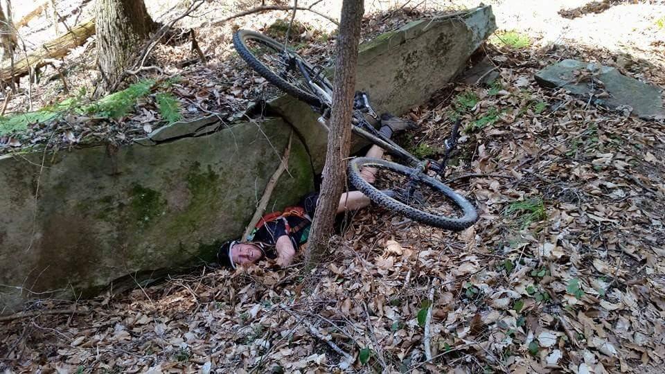 A mountain biker is lying on the ground amidst fallen leaves, partially pinned under a bicycle and a rock. A tree is nearby, and the setting is a wooded area with sunlight filtering through the trees. Cane Creek (sheltowee Trace Trail) mountain bike trail.