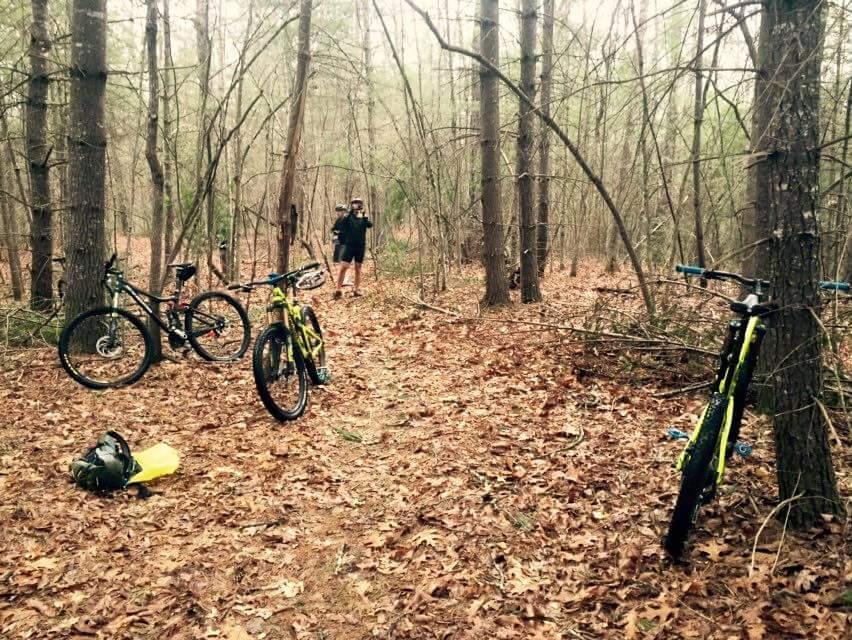 A serene forest scene featuring two mountain bikes placed on a leaf-covered ground. One bike is black and the other is bright yellow, positioned near a backpack. In the background, a person is walking along a dirt path flanked by trees, adding to the outdoor atmosphere. The setting is tranquil, showcasing the beauty of nature and outdoor activities. Big South Fork mountain bike trail.