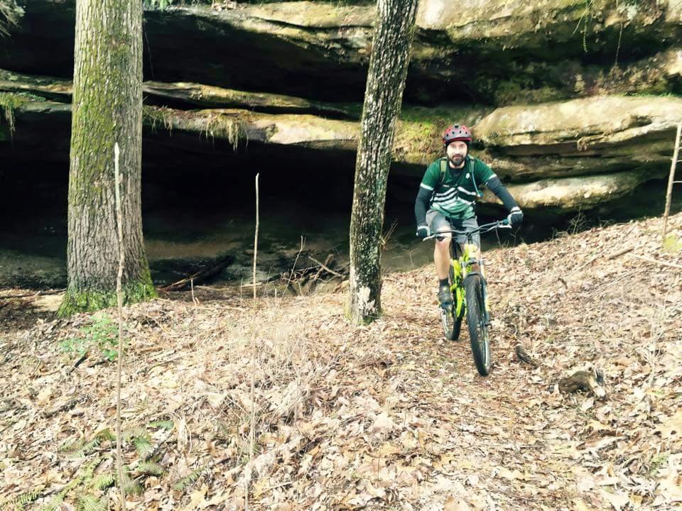 A mountain biker rides along a dirt trail in a forested area, surrounded by tall trees and rugged rock formations. The ground is covered with fallen leaves, and a large rock overhang is visible in the background. The biker, wearing a green shirt and a helmet, navigates the trail with a focused expression. Big South Fork mountain bike trail.