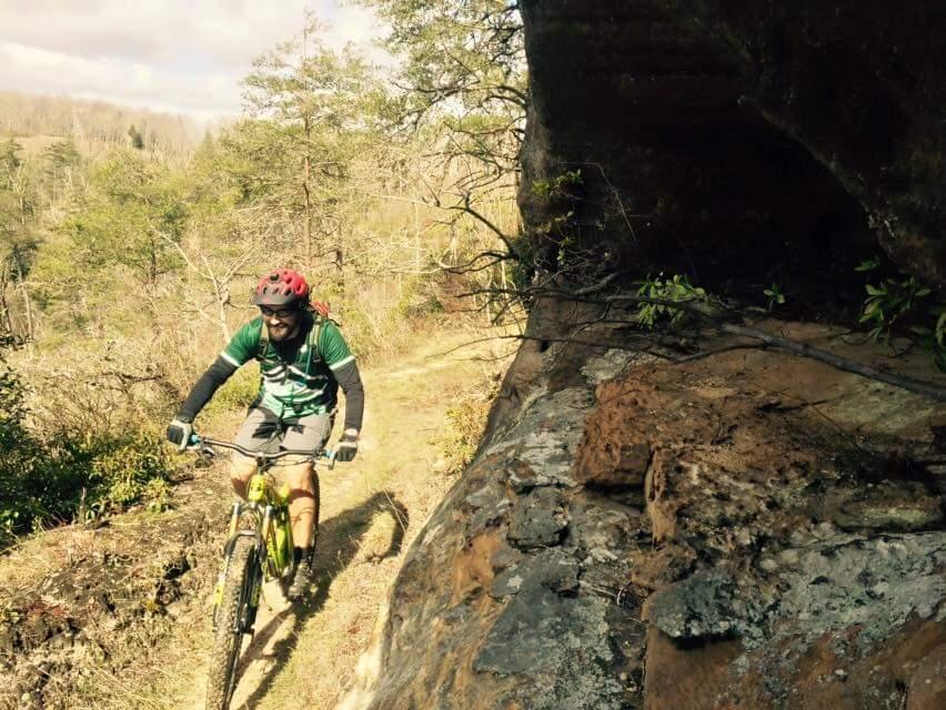 A mountain biker navigates a narrow trail surrounded by lush greenery and rock formations, wearing a red helmet and sports gear. The bike is a bright yellow color, and the cyclist appears focused on the path ahead. The scene is set on a sunny day with a blue sky partially covered by clouds. Big South Fork mountain bike trail.
