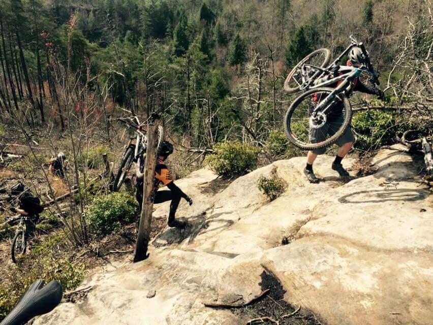 A group of mountain bikers carrying their bikes as they navigate a rocky, steep trail through a forested area, surrounded by trees and underbrush. The terrain appears rugged, and the bikers are focused on their ascent. Big South Fork mountain bike trail.