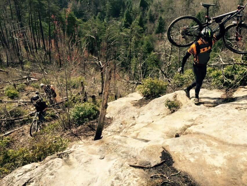 Two mountain bikers navigating a rocky, sloped trail. One cyclist is carrying their bike while climbing up the terrain, and the other is visible further down the slope, riding their bike. The landscape features sparse trees and some greenery, indicative of a rugged outdoor setting. Big South Fork mountain bike trail.