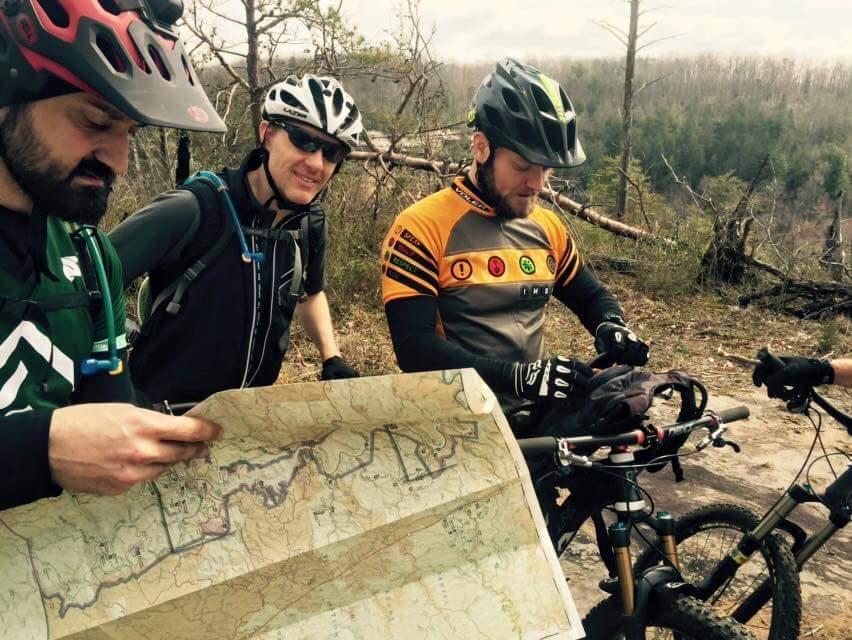 A group of three mountain bikers gathered outdoors, two holding a large map while the third examines a bag on a bike. They are wearing cycling helmets and gear. The background features a forested area with trees and visible trails. Big South Fork mountain bike trail.