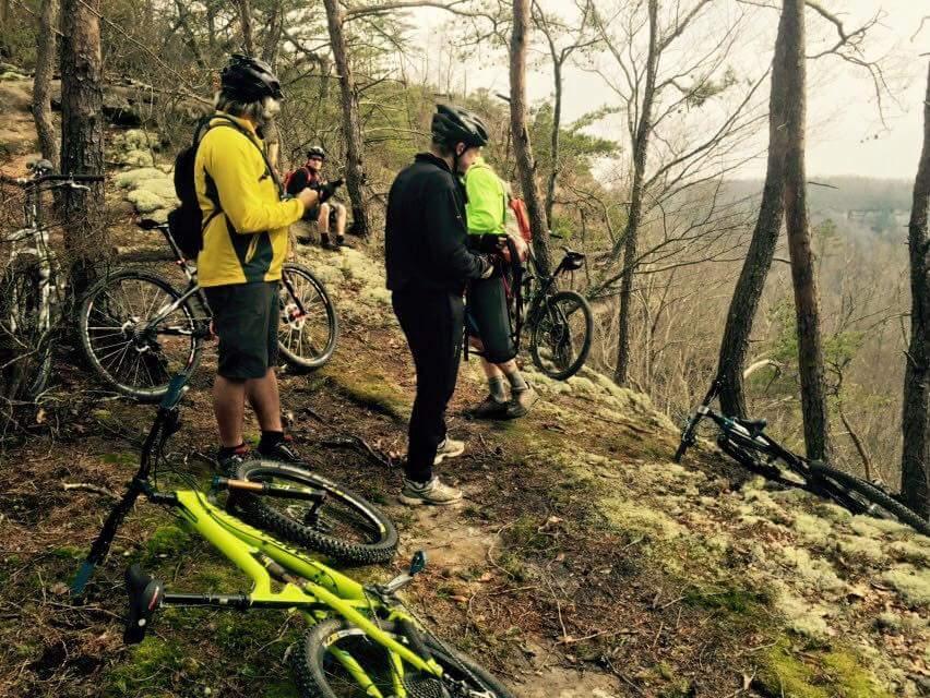 A group of four mountain bikers are gathered on a rocky outcrop surrounded by trees, with two bicycles lying on the ground nearby. The riders are wearing helmets and dressed in outdoor gear. Two of them are looking at their phones, while the others observe the view of the landscape beyond. The scene captures a moment of rest during an outdoor biking adventure. Big South Fork mountain bike trail.