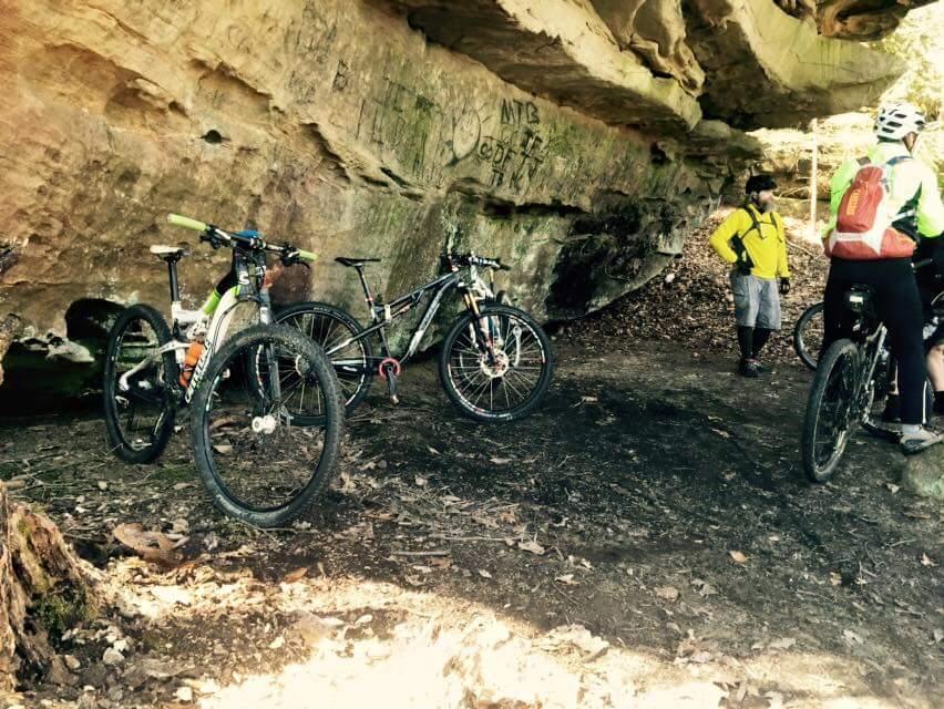 Mountain bikers resting under a rocky overhang, with three bicycles parked nearby. The ground is covered in dirt and leaves, and graffiti is visible on the cave wall. Two riders are discussing while another is adjusting their gear. Big South Fork mountain bike trail.