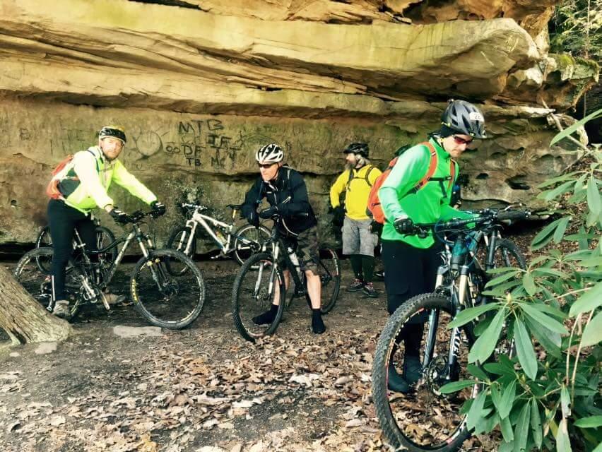 A group of four mountain bikers in a forested area, gathered under a rocky overhang. They are wearing helmets and cycling gear, with two bikes in the foreground and two more in the background. The setting features foliage, rocky textures, and visible graffiti on the rock face. Big South Fork mountain bike trail.