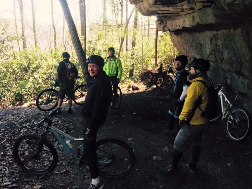 A group of five mountain bikers gathered under a rocky overhang in a wooded area. They are dressed in biking gear and helmets, with bicycles parked nearby. The scene is set in a lush landscape with trees and green foliage, exhibiting a sunny day. One biker is smiling at the camera while others are engaged in conversation. Big South Fork mountain bike trail.