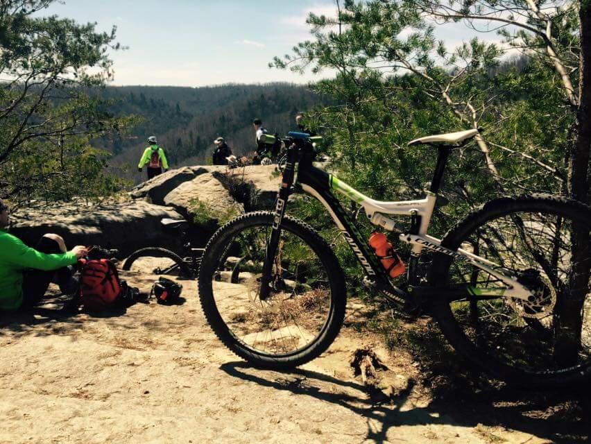 Mountain bikers taking a break on a rocky outcrop with a scenic view in the background. A bicycle is prominently displayed in the foreground, and several people in bright clothing can be seen in the distance enjoying the outdoors. Big South Fork mountain bike trail.