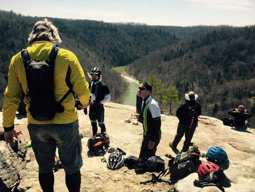 Group of mountain bikers resting on a rocky outcrop with a scenic view of a valley and river below. Some riders are standing and chatting, while others are sitting or preparing their gear. It's a clear day with trees and hills in the background. Big South Fork mountain bike trail.