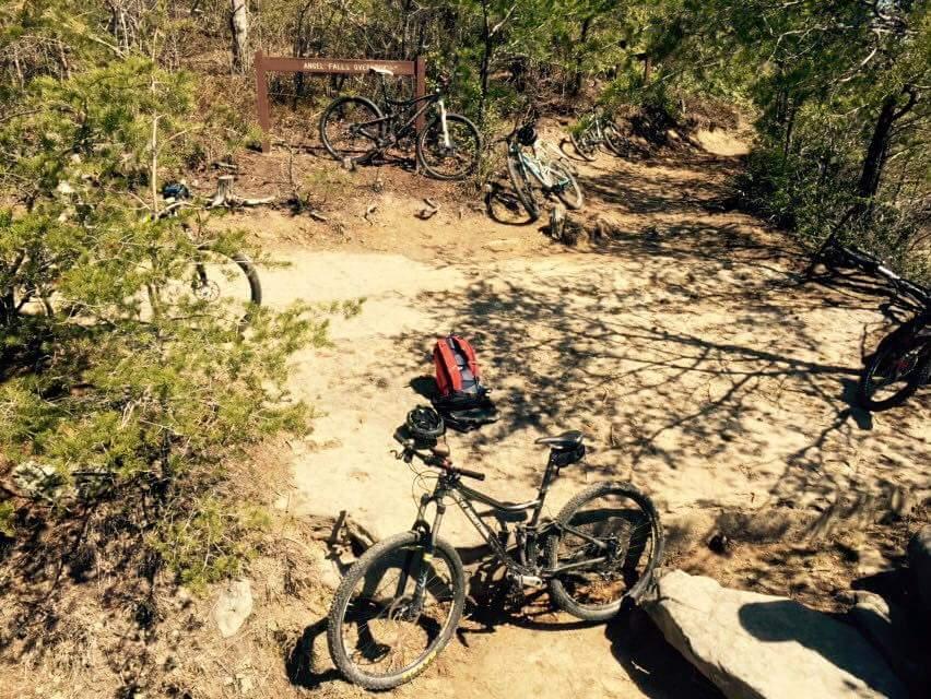 A scenic outdoor trail with several parked mountain bikes near a trail sign labeled "Angels Falls Over." A red backpack is resting on the ground amidst a natural setting featuring greenery, dirt paths, and rocky terrain. Big South Fork mountain bike trail.