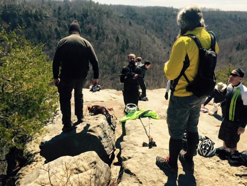 A group of hikers enjoying a panoramic view from a rocky ledge, surrounded by a forested landscape. Some individuals are relaxing while others are gearing up for their hike, with walking poles and backpacks nearby. The scene captures a sunny day outdoors. Big South Fork mountain bike trail.