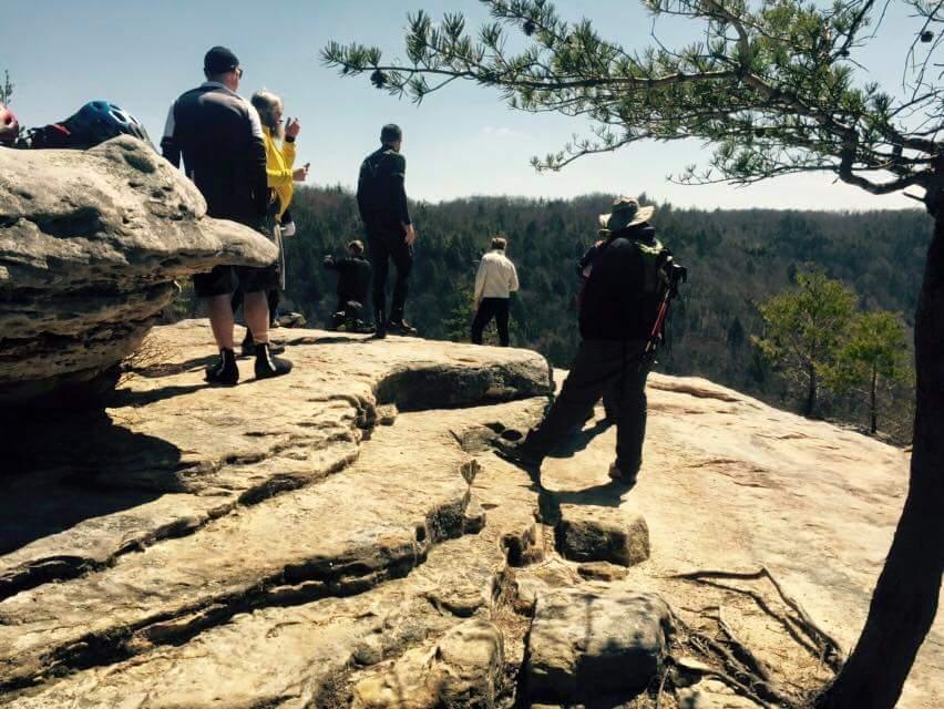 A group of hikers standing on a rocky ledge overlooking a forested valley. Some individuals are taking photos, while others are enjoying the view. The sky is clear and sunny, with a few trees visible nearby. Big South Fork mountain bike trail.