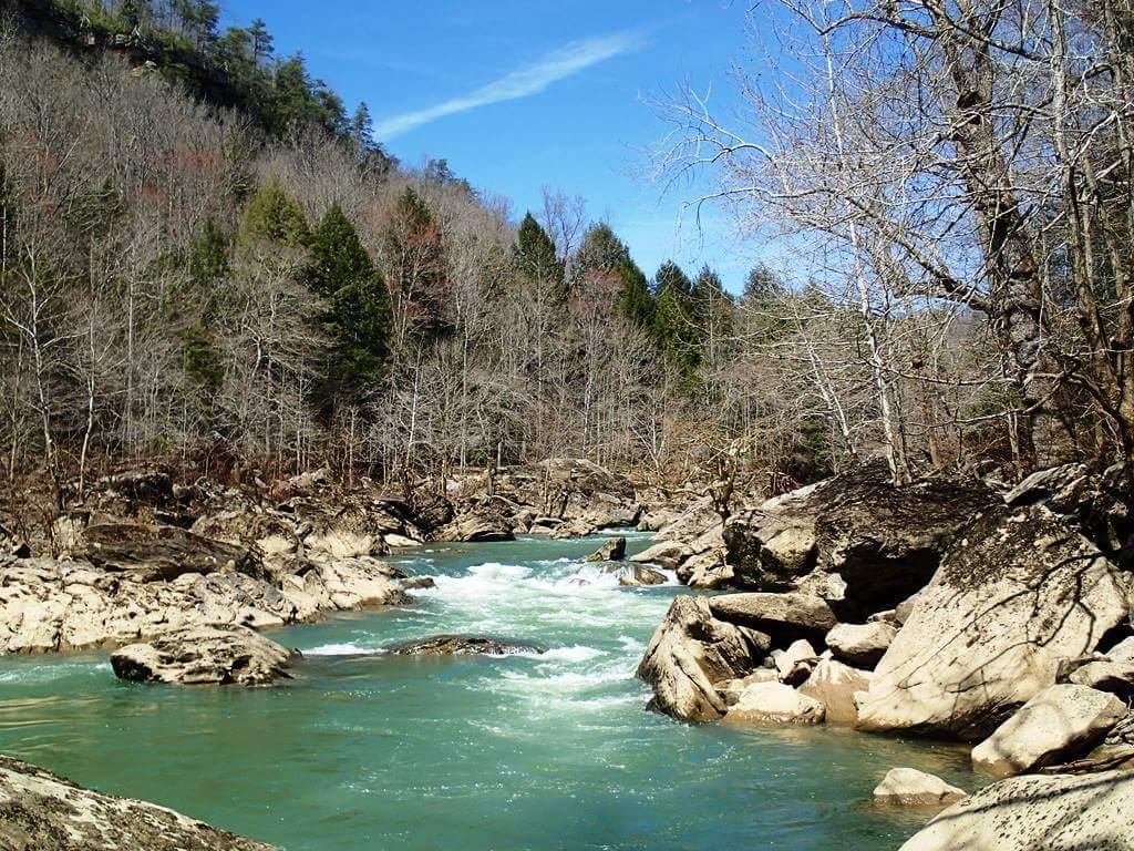 A serene landscape featuring a flowing river surrounded by rocky banks and sparse trees in early spring. The water is a vibrant turquoise color, and the sky is clear with a few wispy clouds. The scene captures the tranquility of nature. Cane Creek (sheltowee Trace Trail) mountain bike trail.