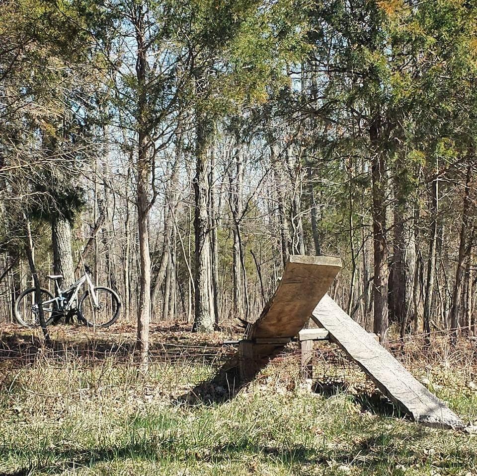 A wooden ramp set up in a forest clearing, with a mountain bike parked nearby. The scene features tall trees with sparse leaves, suggesting early spring. The ramp is placed on a grassy area, surrounded by natural foliage. Skullbuster mountain bike trail.